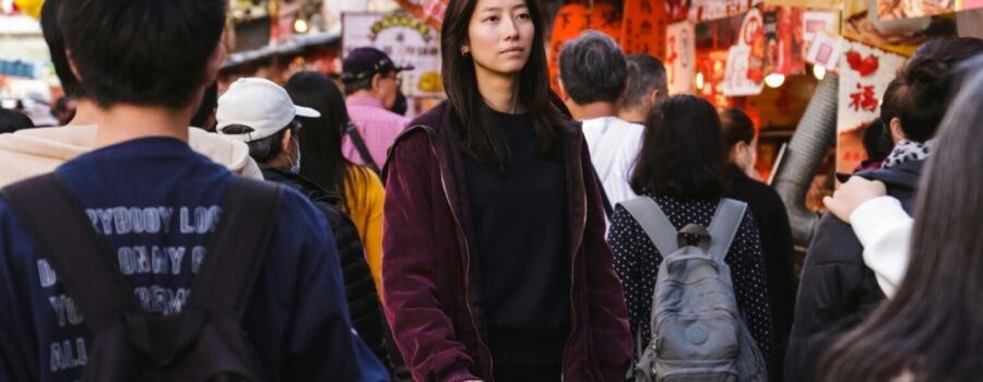 Young woman pushing a silver suitcase through a crowded market street with colorful lanterns and storefronts in the background.