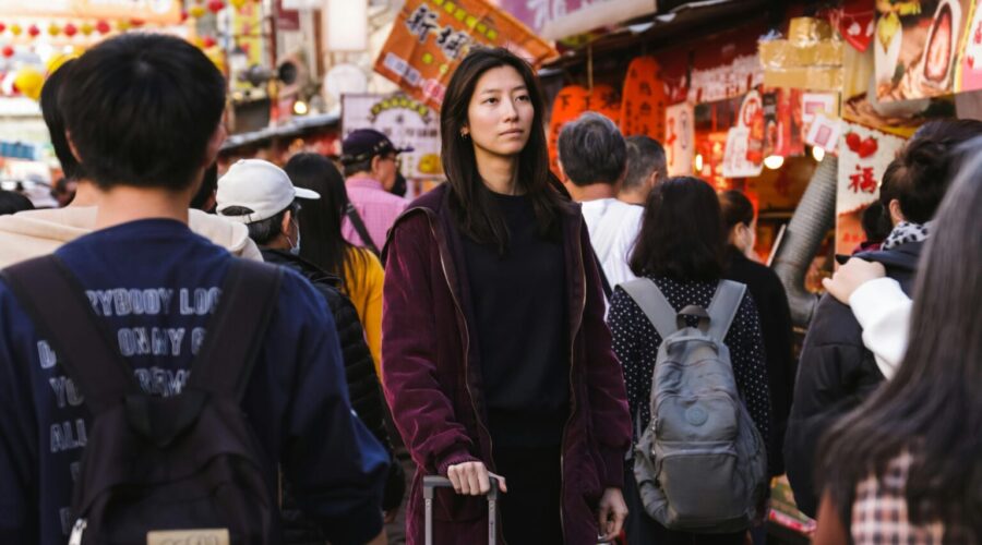 Young woman pushing a silver suitcase through a crowded market street with colorful lanterns and storefronts in the background.