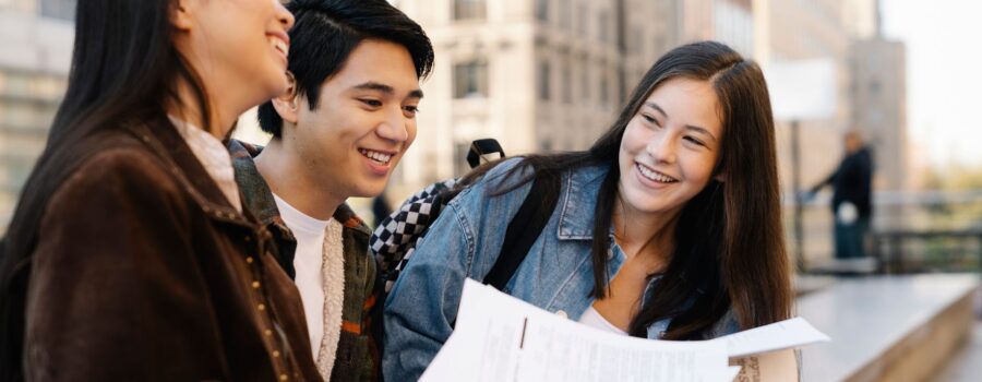 Three smiling friends sit on a bench outdoors, looking at papers together with city buildings in the background.