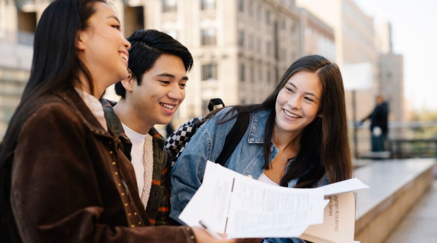 Three smiling friends sit on a bench outdoors, looking at papers together with city buildings in the background.